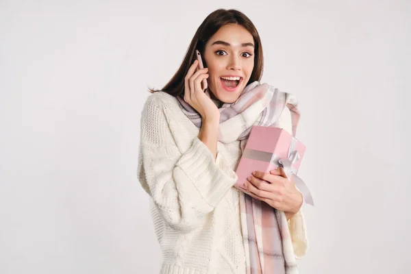 Досить збуджена дівчина в затишному светрі з шарфом тримає подарункову коробку радісно розмовляючи на мобільному телефоні на білому тлі — стокове фото