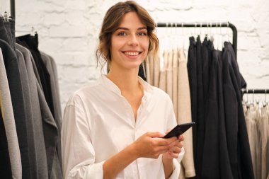 Modern moda galerisindeki cep telefonunu kullanarak kameraya mutlu bir şekilde bakan genç ve güzel bir kadın. 