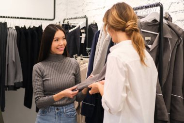 Genç tasarımcı, modern moda galerisinde çalışırken güzel Asyalı kızlara kıyafet seçmekte yardımcı oluyor.