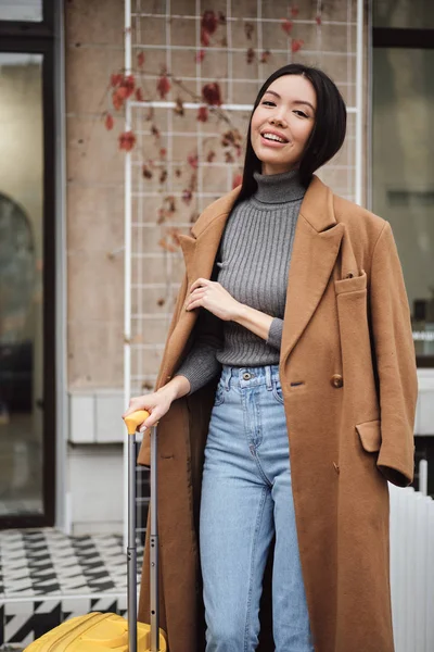 Attractive smiling stylish Asian girl in coat happily looking in camera with yellow suitcase on city street — Stock Photo, Image