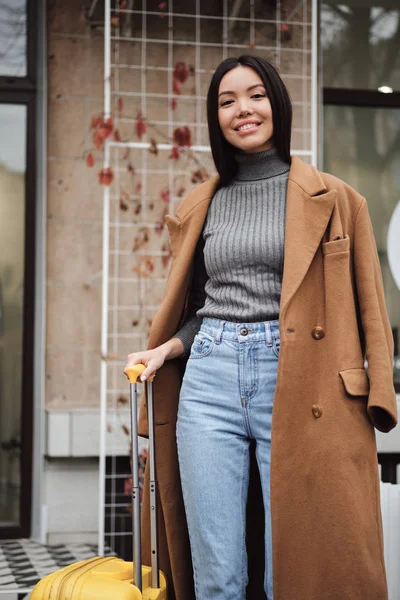 Pretty stylish Asian girl in coat joyfully looking in camera with suitcase on city street — Stock Photo, Image