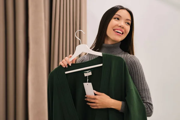 Bastante alegre chica asiática felizmente probándose el cárdigan en la tienda de moda moderna —  Fotos de Stock