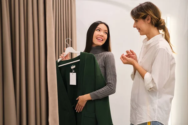 Joven consultor de moda ayudando a probar el cárdigan de la nueva colección de hermosa chica asiática en la tienda moderna —  Fotos de Stock