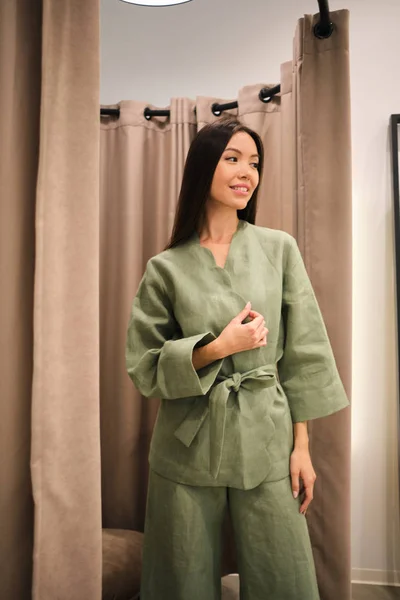 Attractive Asian girl trying on stylish suit dreamily looking away in dressing room of modern clothes store — Stock Photo, Image