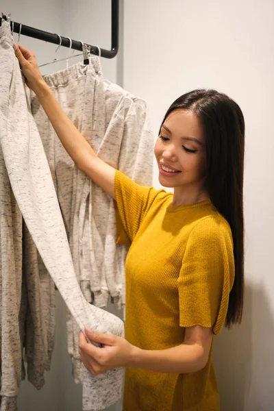 Bastante sonriente chica asiática alegremente elegir ropa en la tienda de moda moderna —  Fotos de Stock