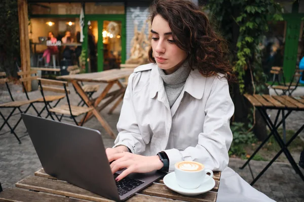 Aantrekkelijk casual brunette meisje in trench jas vol vertrouwen studeren op laptop met koffie in cafe outdoor — Stockfoto