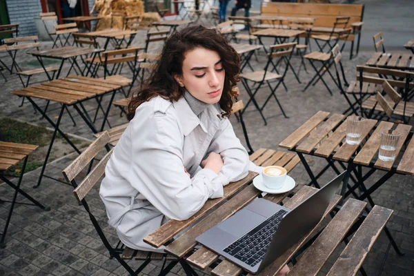 Mooi zelfverzekerd brunette meisje in trench jas werken op laptop met koffie in cafe op straat — Stockfoto