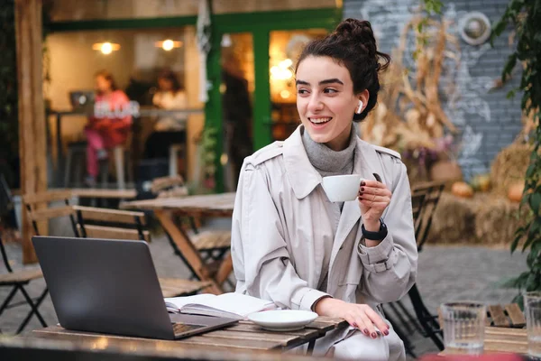 Vrij vrolijk brunette meisje in trench jas gelukkig drinken koffie studeren op laptop in cafe op straat — Stockfoto