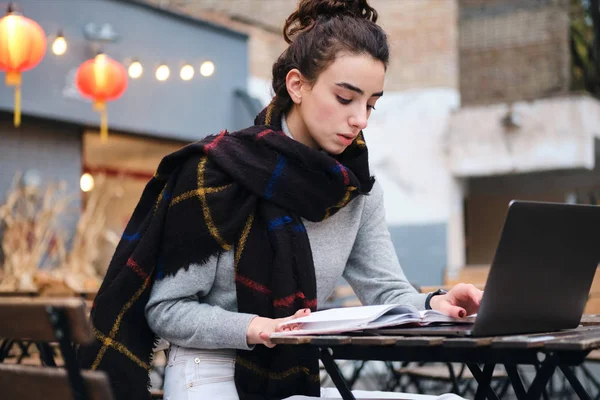 Serious brunette girl with scarf thoughtfully reading book studying with laptop in cafe on street — Stock Photo, Image