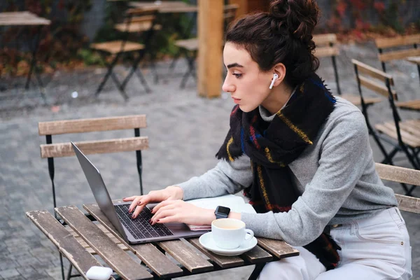 Side view of casual student girl with scarf studying on laptop during coffee break in cafe on street — Stock Photo, Image