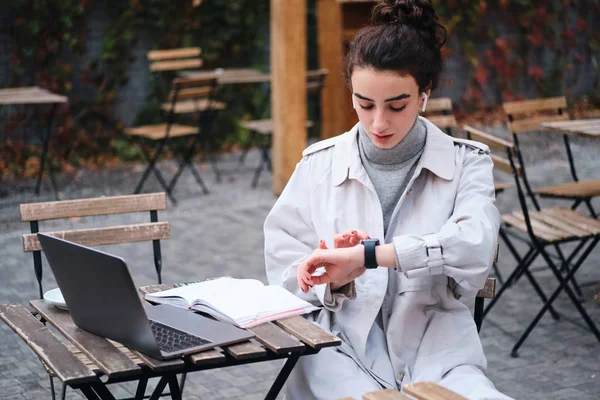 Привлекательная брюнетка в пальто уверенно пользуется умными часами, учится в кафе на улице — стоковое фото