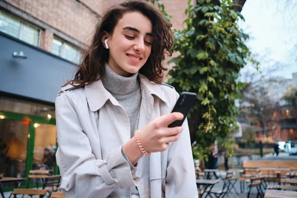 Pretty tersenyum gadis berambut cokelat santai dalam mantel parit gembira menggunakan ponsel di jalan kota — Stok Foto
