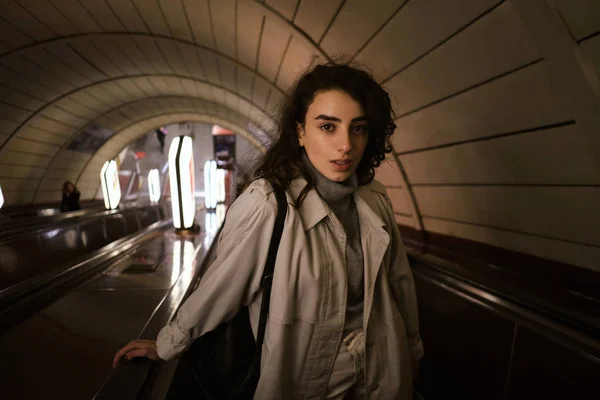 Menina casual atraente em casaco de trincheira olhando atentamente na câmera na escada rolante na estação de metrô — Fotografia de Stock