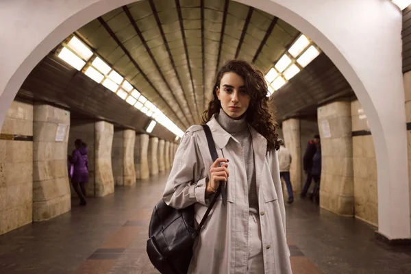 Menina casual atraente em casaco de trincheira com mochila olhando atentamente na câmera na estação de metro — Fotografia de Stock