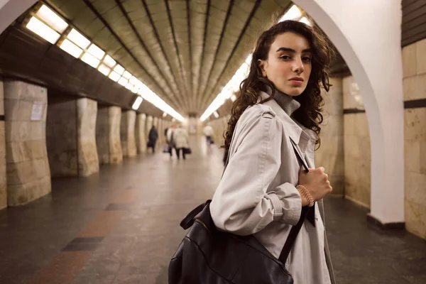 Menina elegante bonita em casaco de trincheira com mochila confiantemente olhando na câmera na estação de metrô — Fotografia de Stock