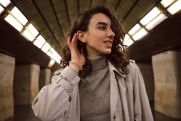 Vista lateral de chica bastante sonriente en gabardina posando alegremente en la estación del metro —  Fotos de Stock