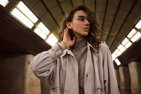 Portrait of attractive casual girl in trench coat sensually looking aside posing at metro station — Stock Photo, Image