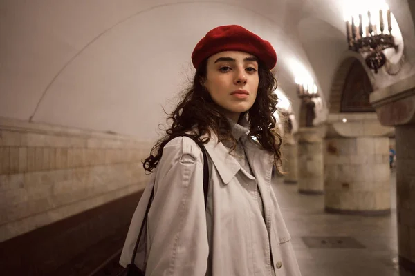 Wspaniała stylowa dziewczyna w płaszczu i czerwony beret z pewnością patrząc w aparacie czeka pociąg na stacji metra — Zdjęcie stockowe