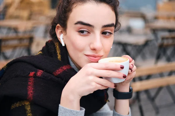 Портрет симпатичной брюнетки с беспроводными наушниками, пьющей кофе тоскливо глядя в сторону — стоковое фото