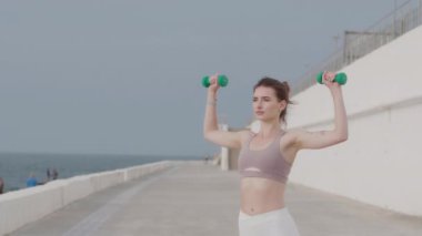 Genç ve çekici bir kadın kumsalda egzersiz yaparken dambıllarla el egzersizi yapıyor.