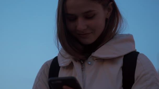 かなりカジュアルな女の子喜んで使用して携帯電話笑顔スタンドアロン屋外で夕暮れ市 — ストック動画