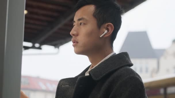 Jonge Aziatische Zakenman Jas Met Draadloze Oortelefoons Bedachtzaam Kijken Naar — Stockvideo