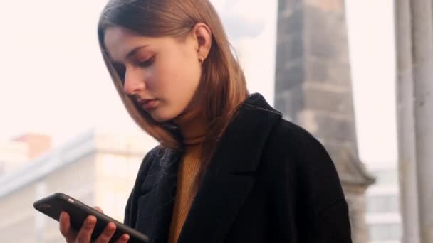 穿着外套的漂亮女孩专心致志地使用手机穿过城市街道的侧视图 — 图库视频影像