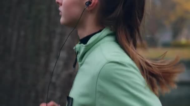 川沿いの秋の都市公園で思慮深く走るイヤホンの美しいスポーティーな女の子の側面図 — ストック動画