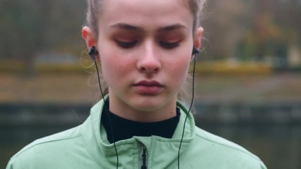 Portrét krásné sportovní dívka ve sluchátkách sebevědomě dívá do kamery po spuštění v podzimním městském parku na břehu řeky