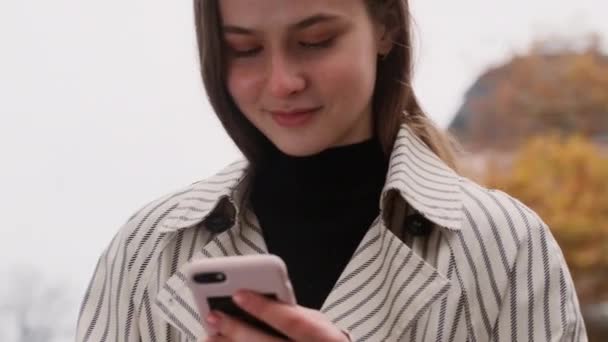 用手机在城市街道上愉快地等待着穿着风衣的漂亮女孩的近照 — 图库视频影像