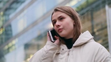 Ceketli çekici bir kız cep telefonuyla konuşuyor. Şehir sokağına dikkatlice bakıyor.