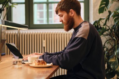 Kafede kahve molası sırasında dizüstü bilgisayarda çalışan çekici sakallı adamın yan görüntüsü.