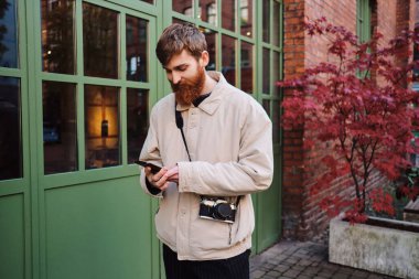 Genç, sakallı bir adam cep telefonunu açık havada kullanıyor.