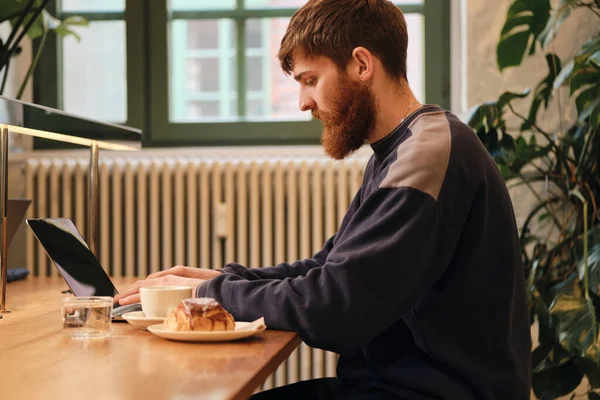 Seitenansicht eines jungen attraktiven bärtigen Mannes, der während der Kaffeepause im Café am Laptop arbeitet — Stockfoto
