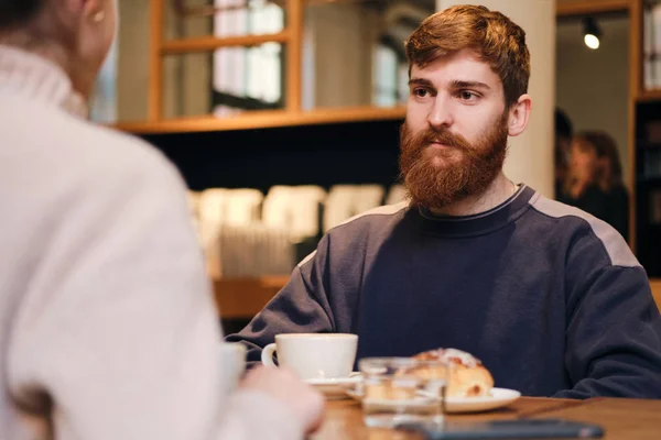 Молодой привлекательный бородатый мужчина пристально смотрит на девушку во время кофе-брейка в кафе — стоковое фото