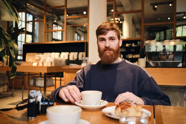 Young attractive bearded man intently looking in camera having coffee break in modern cafe — 스톡 사진