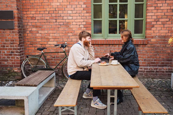 Joven pareja elegante felizmente trabajando juntos en el ordenador portátil durante el descanso de café en la cafetería de la calle — Foto de Stock