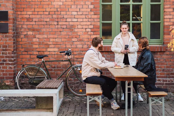Jóvenes amigos elegantes felizmente hablando y tomando café descansando juntos en la cafetería en la calle — Foto de Stock