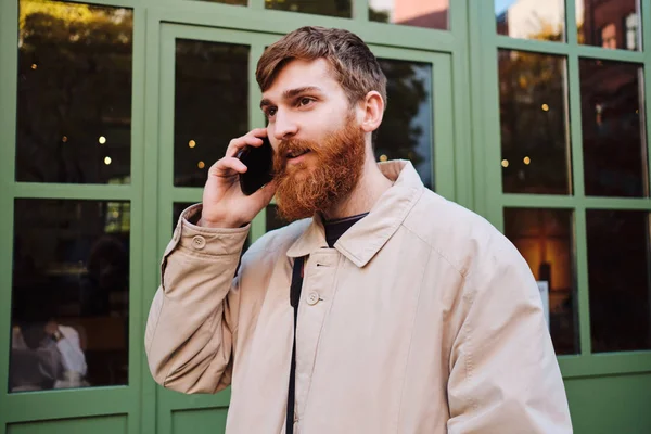 Young handsome bearded man dreamily looking away talking on cellphone outdoor — 스톡 사진