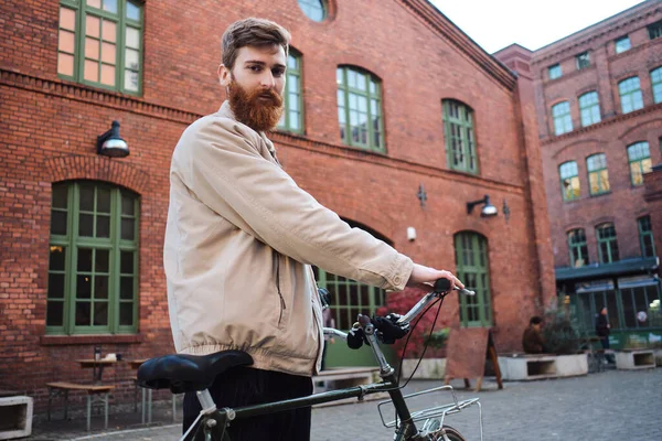 Young attractive bearded man with bicycle confidently looking in camera on street — Stock Photo, Image