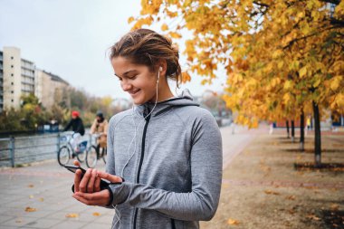 Kulaklıklı neşeli kızın portresi mutlu bir şekilde parkta koşarken cep telefonu hazırlıyor.