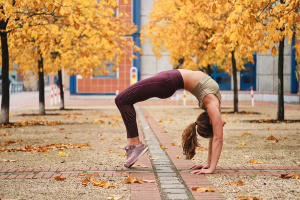 Joven chica deportiva atractiva haciendo ejercicio de puente durante la práctica de yoga en la calle de otoño —  Fotos de Stock