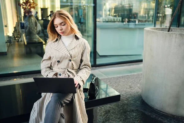 Young stylish businesswoman thoughtfully working on laptop sitting on city street alone — 스톡 사진