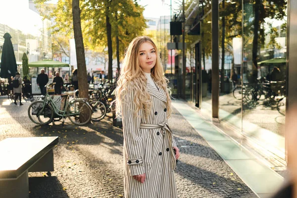 Piękna blondynka w stylowym płaszczu spaceru przez przytulną ulicę miasta w słoneczny dzień — Zdjęcie stockowe
