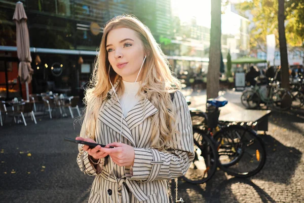 Vakker blond jente lytter til musikk i øretelefoner på mobiltelefon og koser seg i byens gate – stockfoto