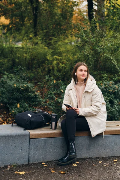 Aantrekkelijk casual meisje gelukkig luisteren muziek op mobiele telefoon rusten op bank in park — Stockfoto