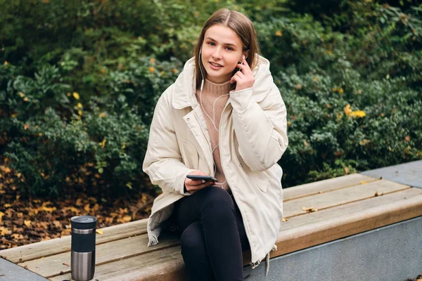 Atrakcyjny casual dziewczyna szczęśliwie patrząc w aparacie słuchania muzyki na komórkę w parku — Zdjęcie stockowe