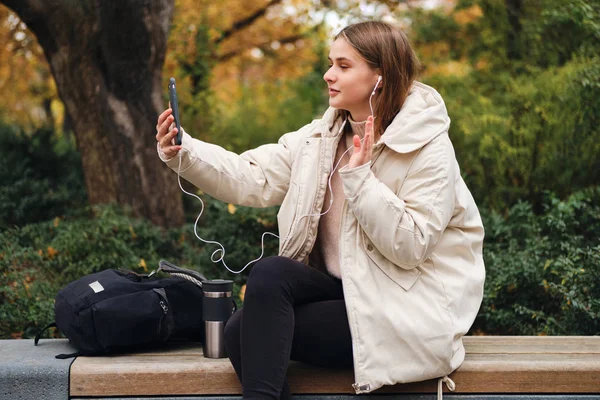 Kaunis tyttö untuvatakki heiluttaa hi ele puhuu video chat matkapuhelin puistossa — kuvapankkivalokuva