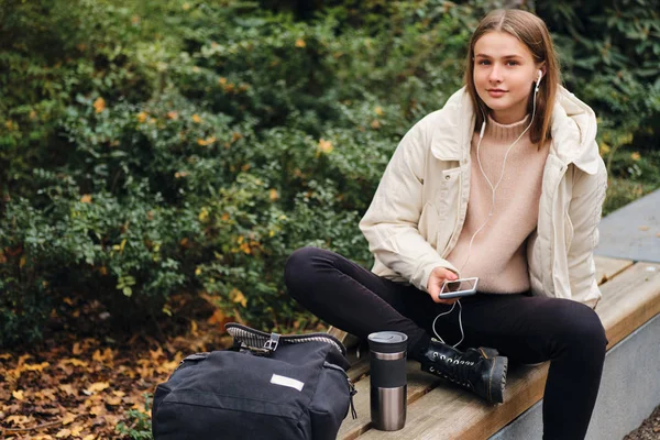 Menina muito sorridente sonhadoramente ouvir música no celular descansando no banco no parque da cidade — Fotografia de Stock