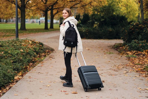 Attraktives lächelndes Mädchen in Daunenjacke mit Koffer, das freudig in die Kamera schaut, wenn es im Park spaziert — Stockfoto